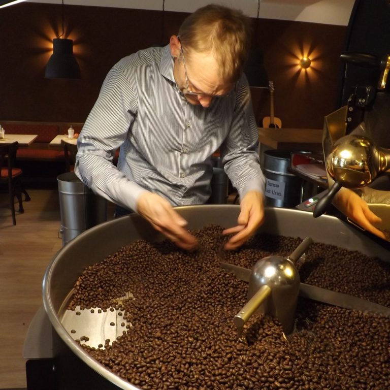 Ein Mann mit Brille inspiziert geröstete Kaffeebohnen in einem großen Metallbehälter in einer gemütlichen Café-Atmosphäre. Im Hintergrund si